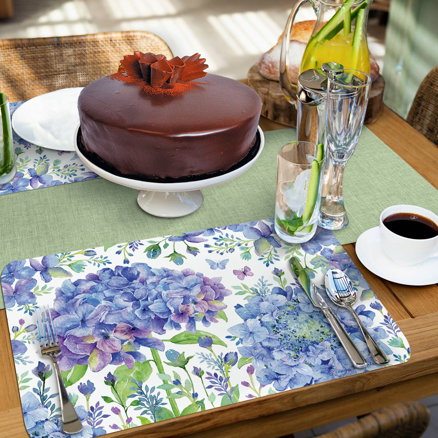 A table setting featuring a delectable chocolate cake, refreshing beverage, and easy-care Beautiful Hydrangea Reversible Rectangular Plastic Placemats. These BPA-free placemats ensure peace of mind while beautifully enhancing the floral theme with their blue hydrangea design.