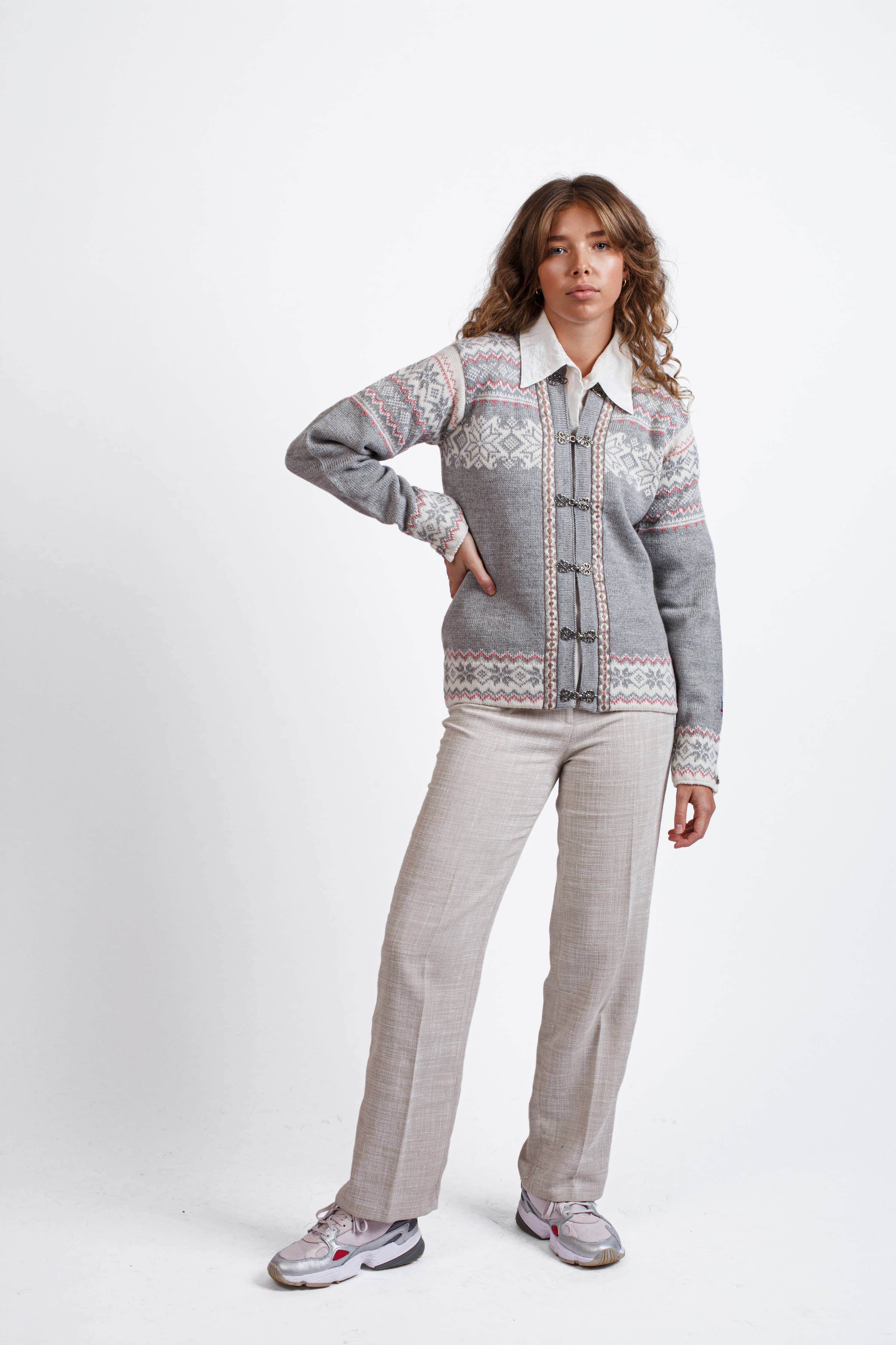 A person stands against a plain background, wearing the Voss Wool Cardigan in grey by Norlender, crafted from pure new wool. Light pants and sneakers complete the look. With hands relaxed by their side, one slightly on the hip, they embody a casually confident style.