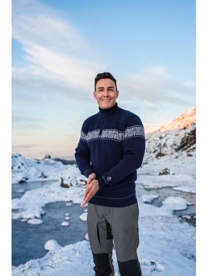 Amidst a snowy landscape with mountains in the background under a clear sky, a person smiles wearing the elegant Breheimen Jutunheimen Merino Wool Zip Cardigan in navy, exemplifying Nordic design.