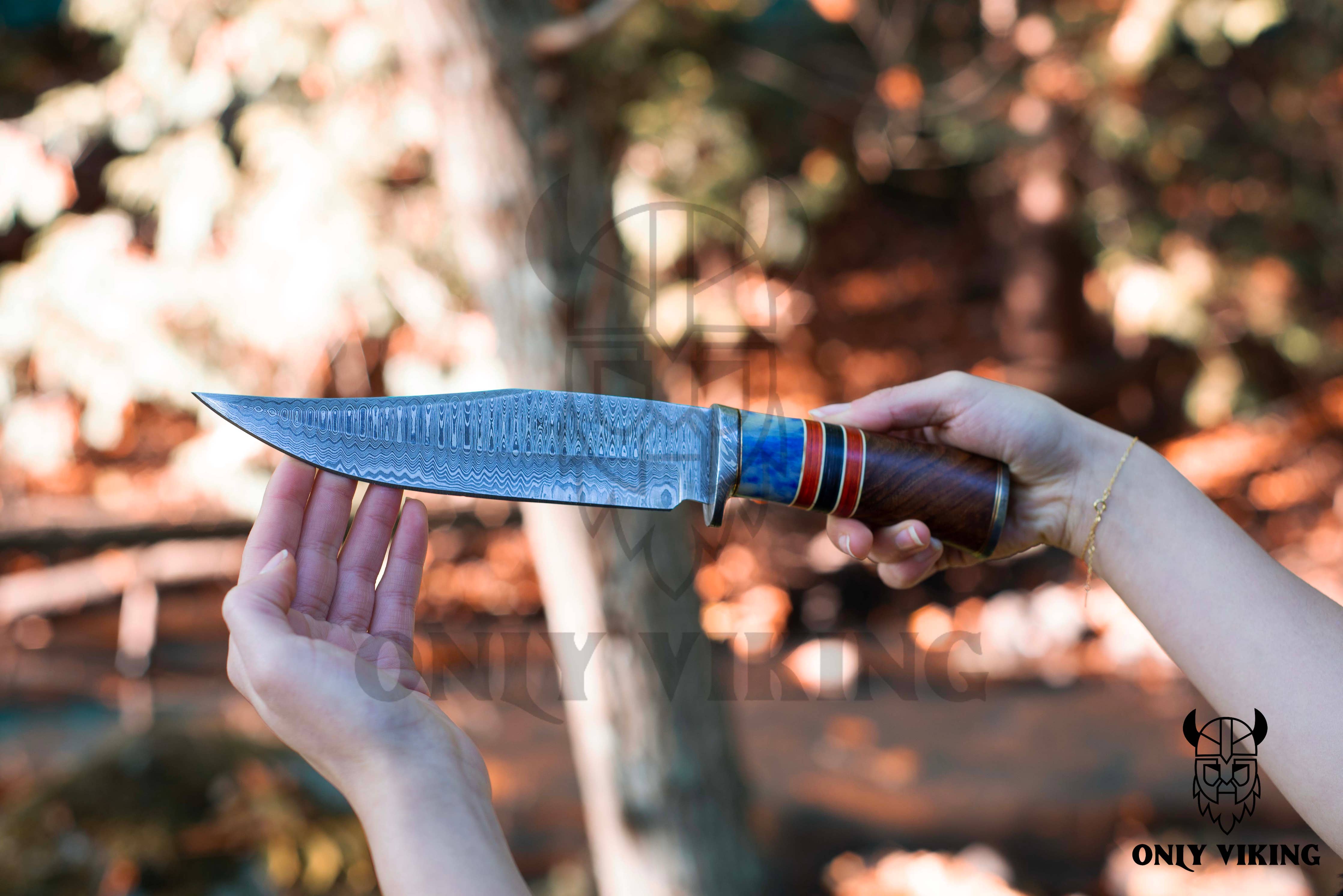 A person holds a Damascus Steel Hunting Knife, with its patterned blade gleaming and wooden handle firm in their grip, set against a blurred outdoor background.