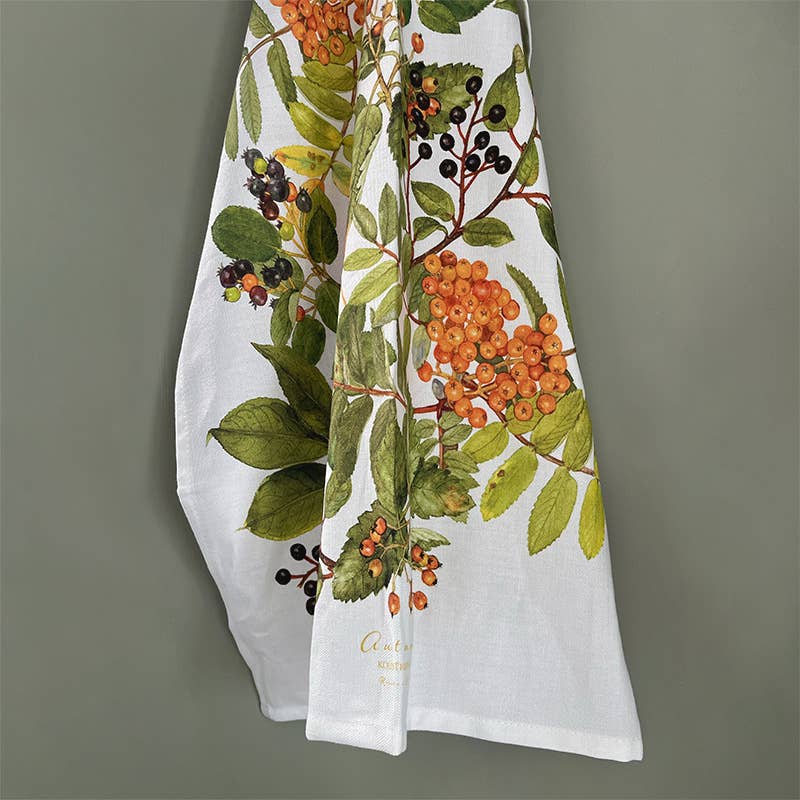 The Tea Towel: Autumn by Kirsten Tind Koustrup & Co, crafted from 100% organic cotton and adorned with a botanical design of green leaves and orange berries, elegantly enhances the kitchen decor when displayed on a light green wall.