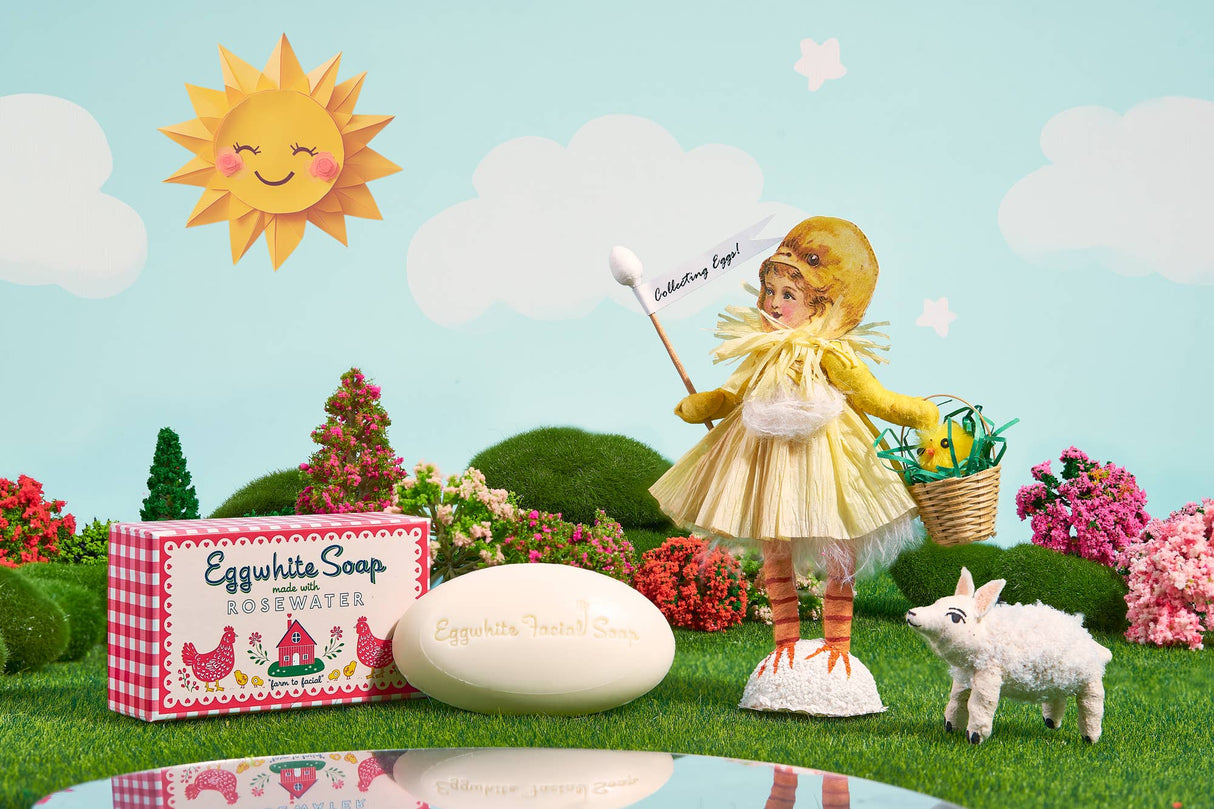 A whimsical scene with an Eggwhite & Rosewater Facial Soap 1.76oz box, oval soap bar, doll in a chick costume holding a flag, small lamb figure, and decorative trees under a sunny sky—perfect for radiant skin.