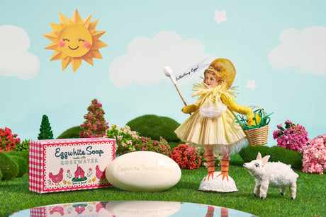 A whimsical scene with an Eggwhite & Rosewater Facial Soap 1.76oz box, oval soap bar, doll in a chick costume holding a flag, small lamb figure, and decorative trees under a sunny sky—perfect for radiant skin.