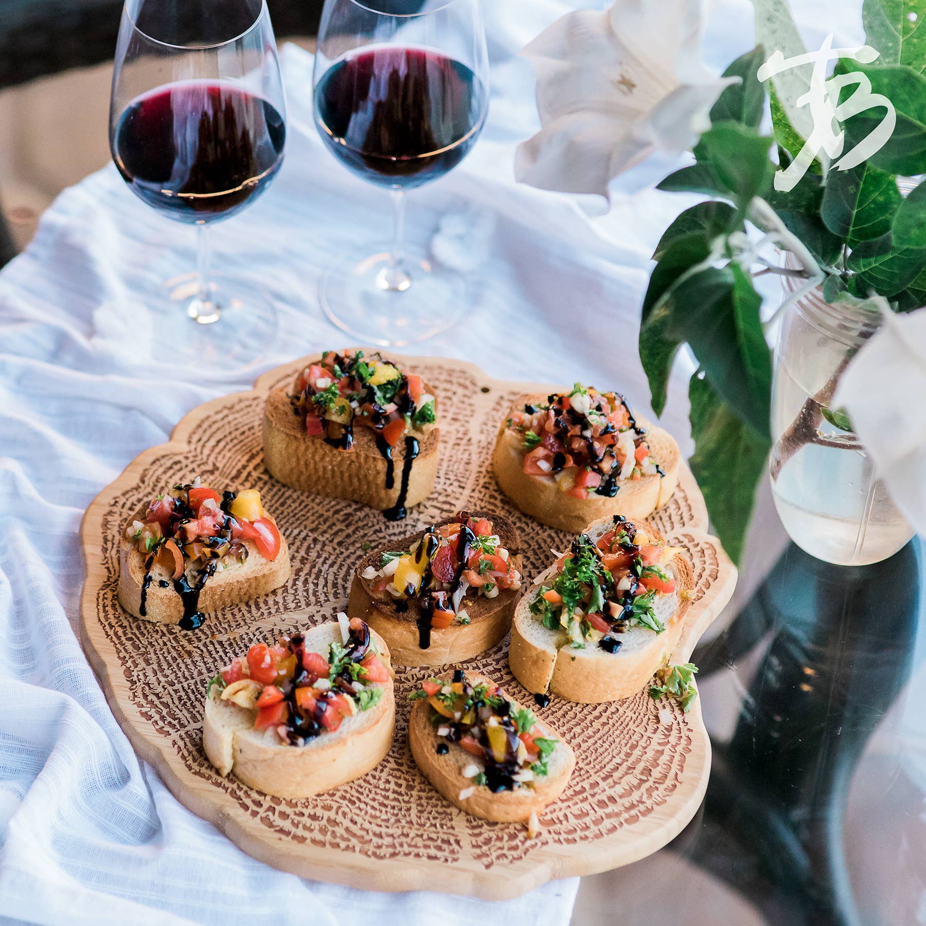 A Serving Board: Tree Of Life 13" elegantly presents bruschetta topped with colorful vegetables, paired with two glasses of red wine and a vase containing green leaves. The intricate engraved artwork on this sustainable bamboo piece enhances the artistic ambiance of the setting.