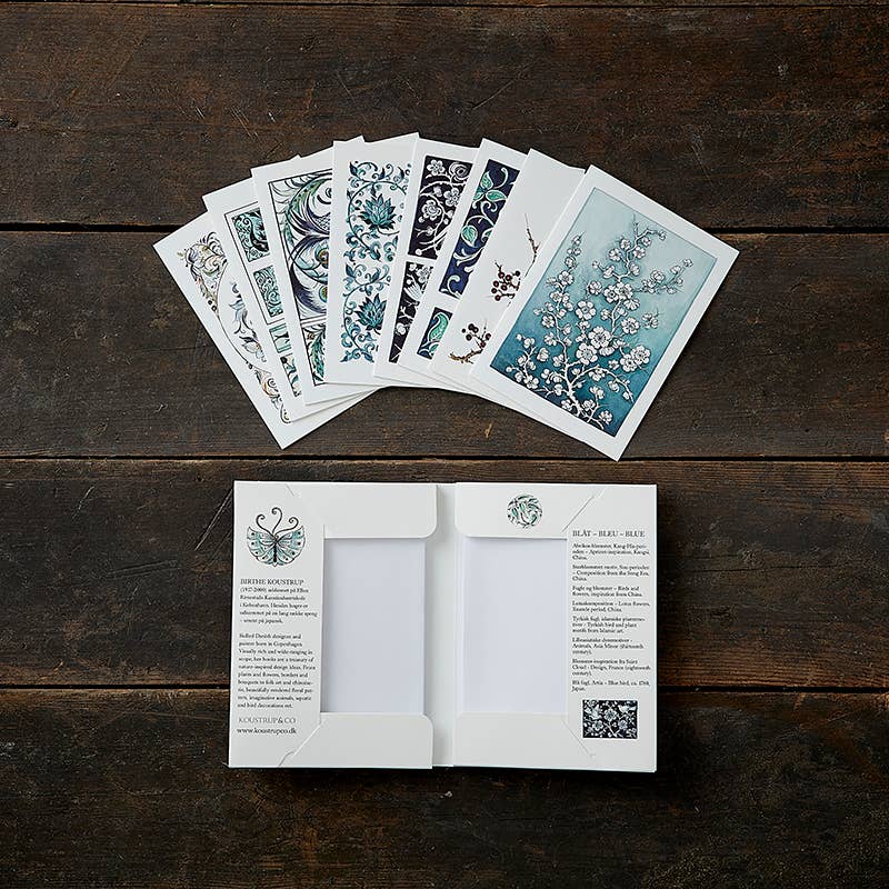 A set of eight "Blue in Blue" cards, designed by Birthe Koustrup and featuring blue motifs, is elegantly arranged on a wooden surface alongside a floral design booklet. These exquisite items are crafted from environmentally certified FSC paper and come complete with envelopes.