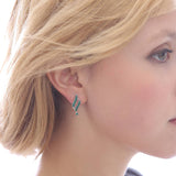 Close-up of a woman with short blonde hair wearing April Crystal Swarovski® Crystals Spiral Dangle Earrings in green on her left ear, facing right against a bright background.