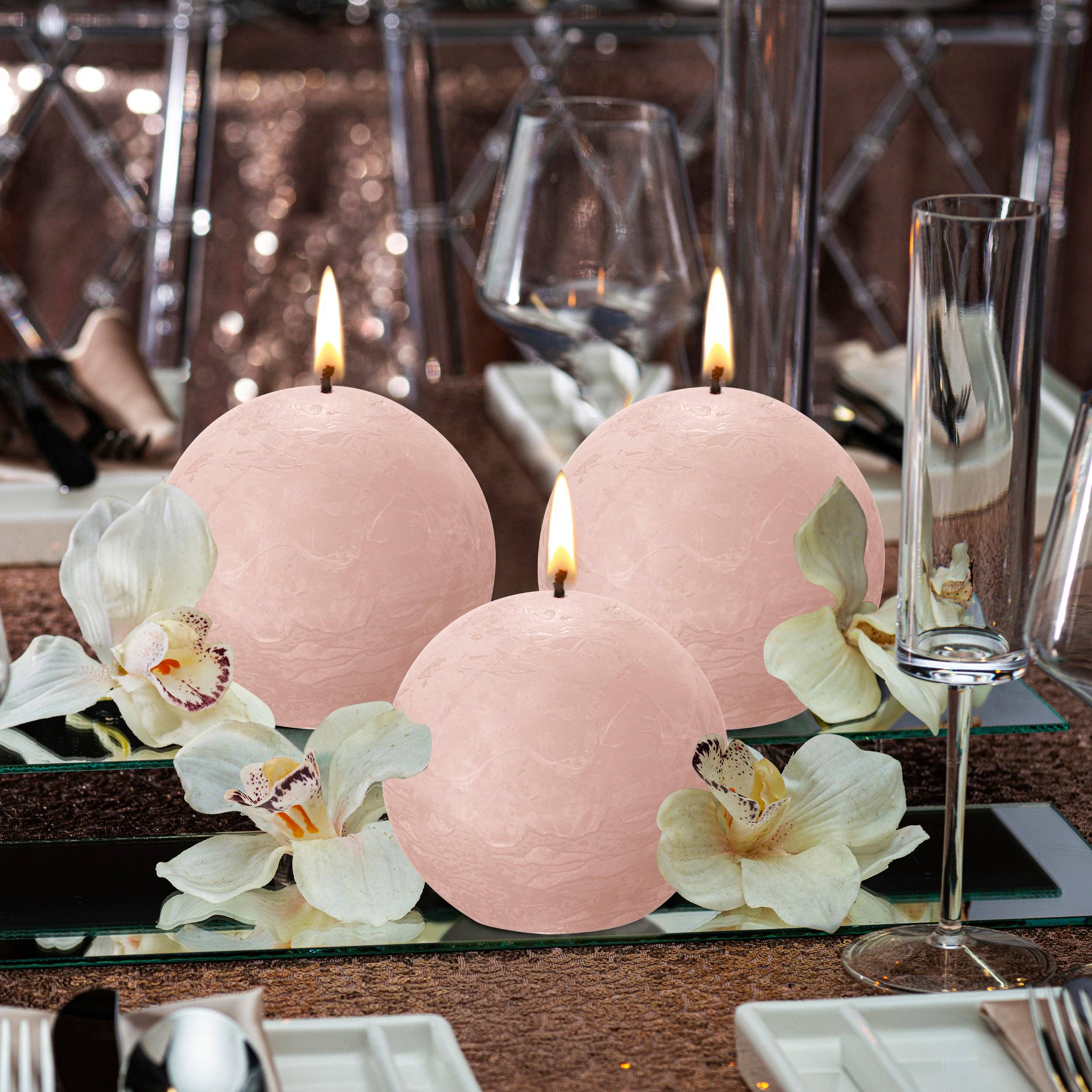 A lit Misty Pink Rustic 3" Ball Candle sits on a mirrored surface with white orchids, surrounded by elegant glassware and table settings, highlighting its soft pink hue and plant-based wax craftsmanship.