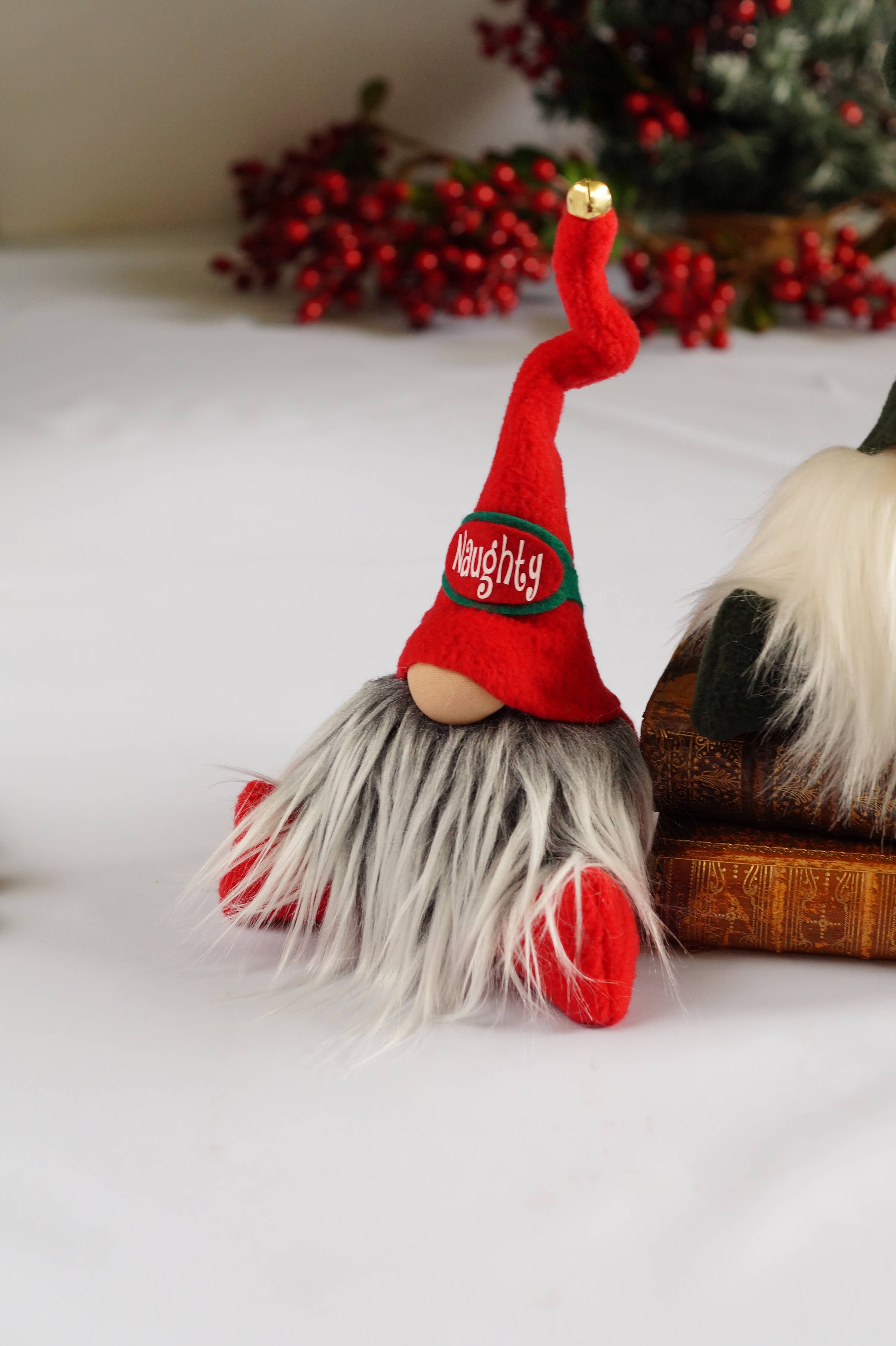 The Naughty Christmas Gnome, a small figurine featuring a red hat, is perched on a white surface beside a wooden block. Festive red berries adorn the background, adding playful warmth ideal for cozy holiday gatherings.