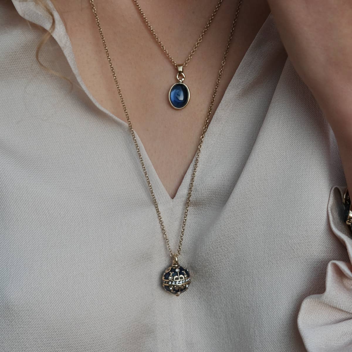 A person is wearing two necklaces: the A&C Oslo Gold Plated Glass and Crystal Pendant Necklace with a blue oval crystal, and another featuring a round ornate design, both paired with a light-colored blouse.