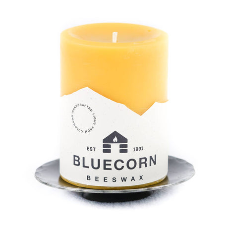 A yellow beeswax candle labeled Bluecorn Beeswax sits on a Forged Steel Pillar Candle Base (4.75" diameter) against a white background.