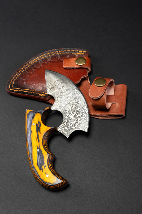 The Knife: Alaskan Ulu Style Blade features a high carbon Damascus steel patterned blade and a yellow-black wooden handle, shown beside its brown leather sheath on a black surface.