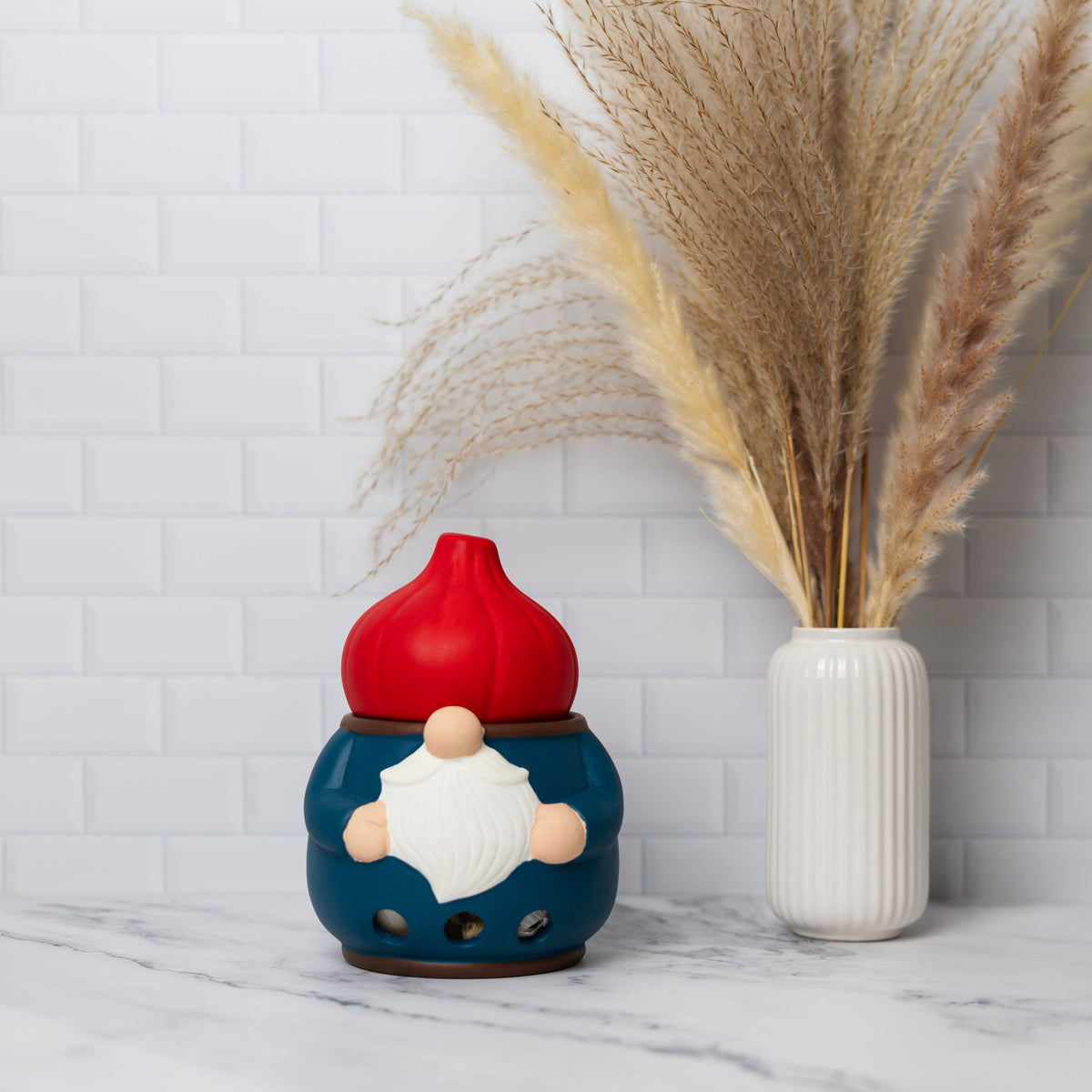 Ceramic gnome garlic keeper next to a white vase filled with dried pampas grass, both placed in front of a white tile wall.