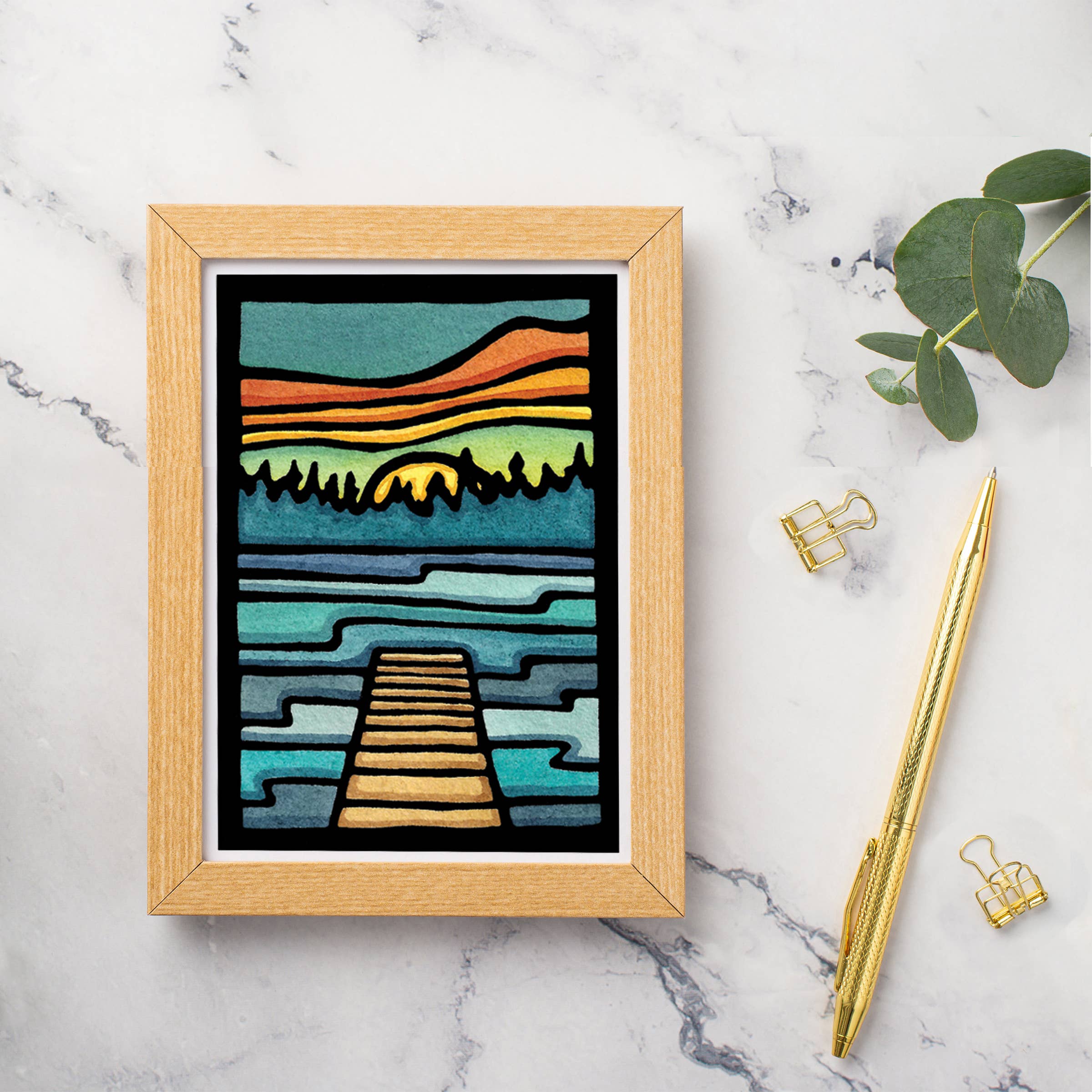 Greeting card featuring the "Lake Life Dock" by Sarah Angst, depicting a sunset over a lake with a wooden dock, paired with a marble surface, gold pen, and binder clips.