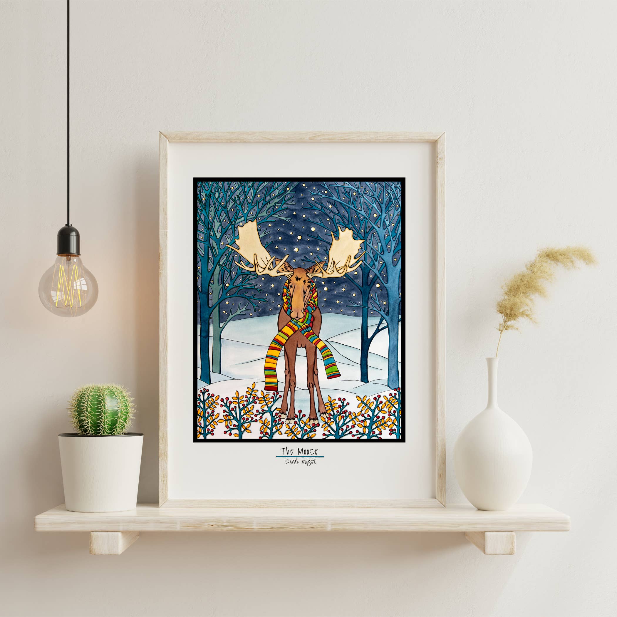 An 8"x10" giclee print titled "Artwork: Holiday Moose" captures a moose adorned with a scarf, nestled in a snowy forest scene. Displayed elegantly on a shelf, this framed illustration is surrounded by Christmas decorations including a plant, vase, and illuminated bulb that enhance the festive atmosphere.