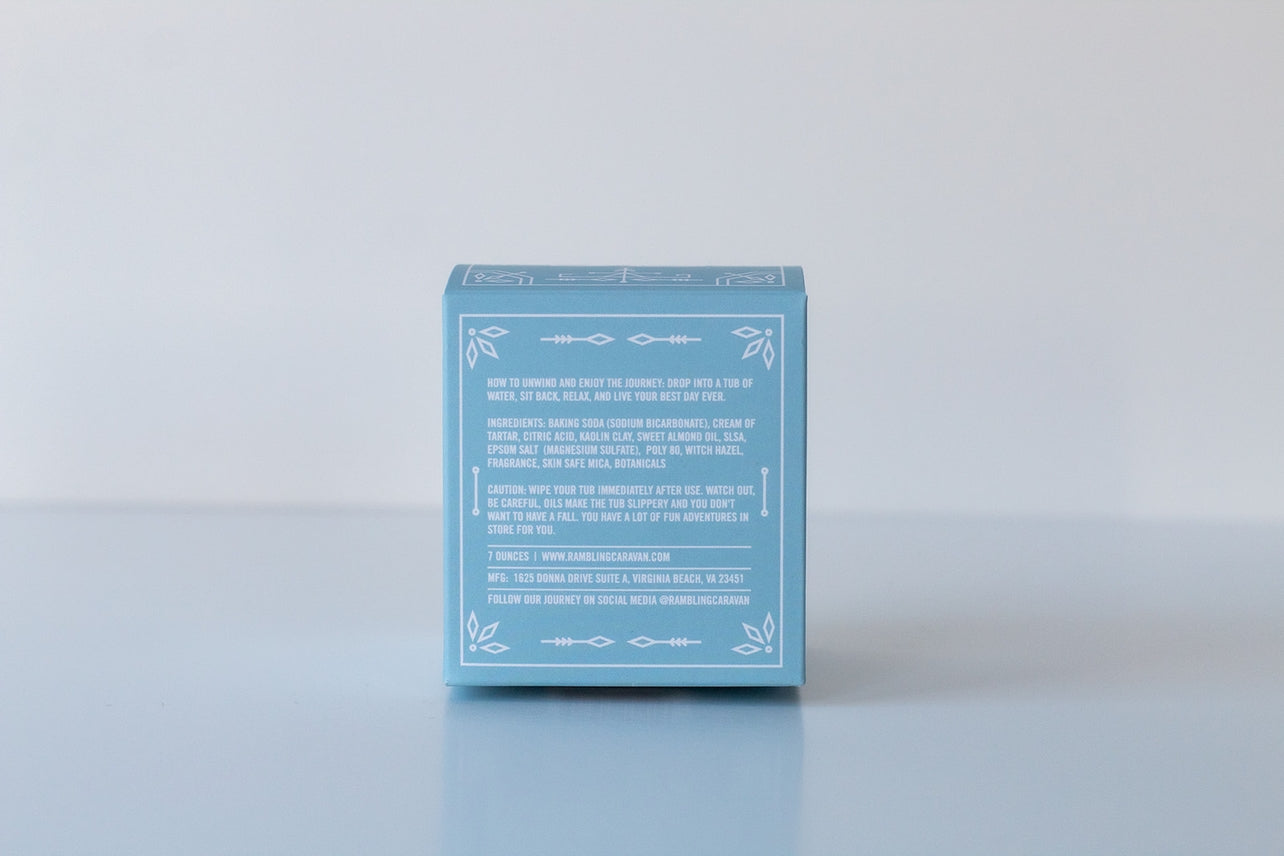 A blue box labeled Bath Bomb: Coastal Vibes displays white text with ingredients, skin-care instructions, and company details for this luxurious handmade-in-USA bath bomb, set on a light surface against a neutral background.