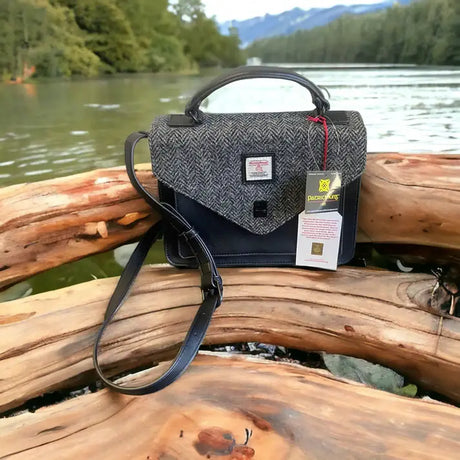 A Harris Tweed Large Satchel - Grey with a shoulder strap and tags hangs on a smooth log by a river, with trees and mountains in the background.