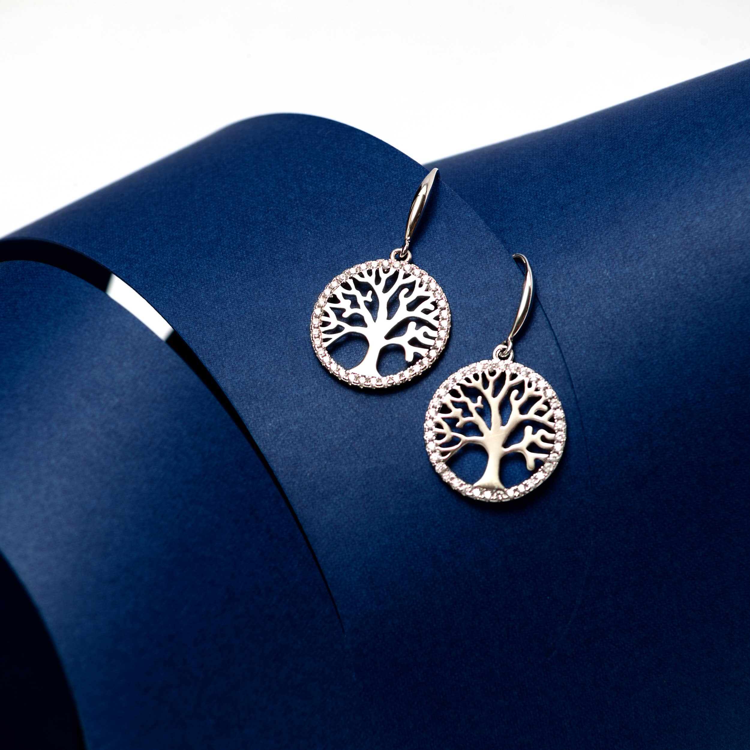 A pair of Tree of Life in Sterling Silver earrings with beautiful luster, featuring delicate tree designs and Cubic Zirconia accents, displayed on a curved dark blue surface.