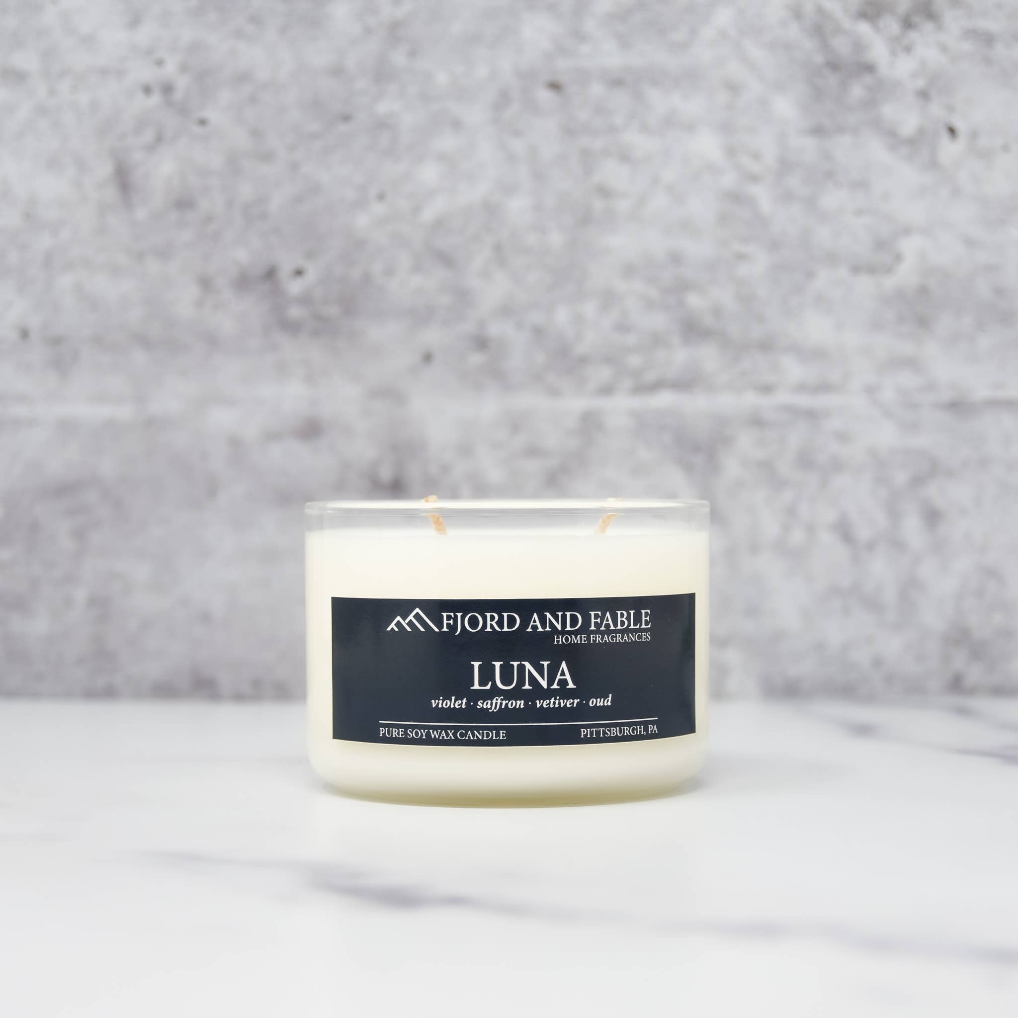 A 4oz Luna Candle by Fjord and Fable, made of white soy wax in a glass container, is displayed on a white surface with a gray stone background.