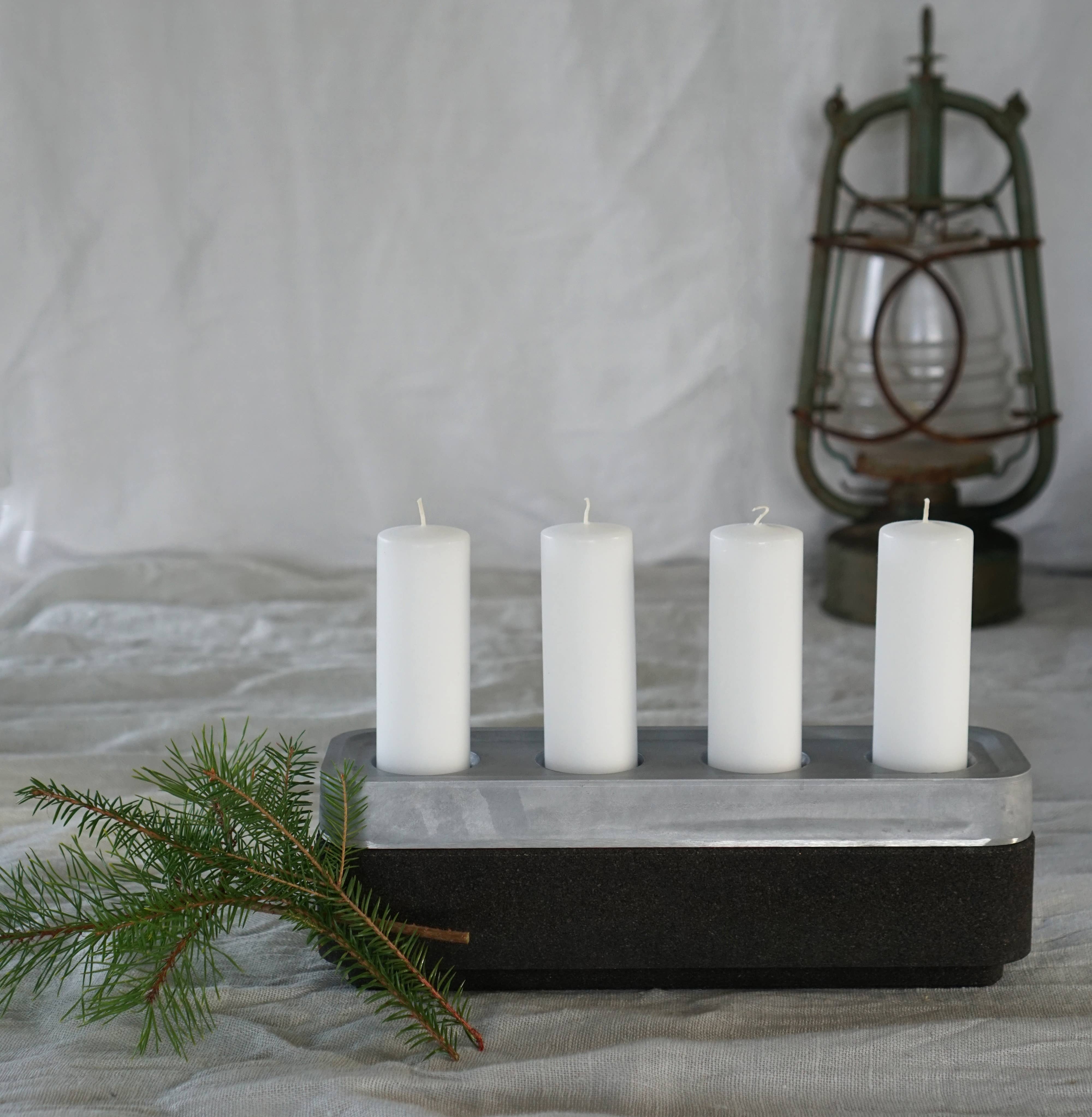 Four white pillar candles sit on a rectangular Stumpastaken holder with a nearby small evergreen branch and antique lantern. The scene exudes eco-friendliness, featuring the Candle Holder: Stumpastaken Corkbowl Black crafted from recycled cork for added texture and warmth.