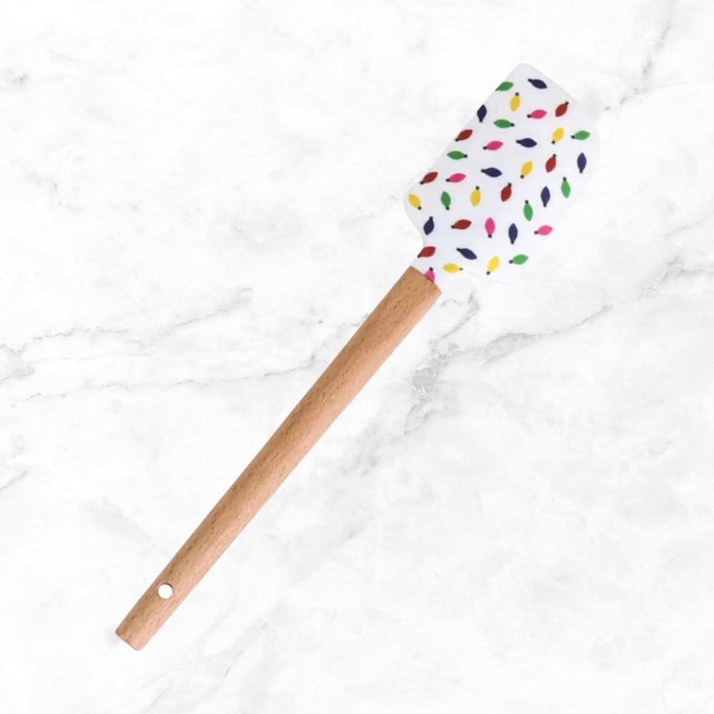 A silicone spatula with a wooden handle and a white head decorated with colorful Christmas lights, resting on a white marble surface.