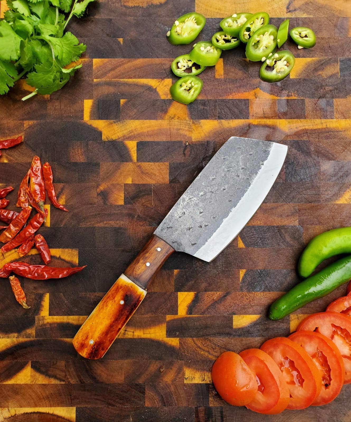 The Handmade Carbon Steel Cleaver for Kitchen & Outdoor Use rests on a wooden cutting board, surrounded by chopped jalapeños, sliced tomatoes, dried red chilies, green chili peppers, and cilantro.