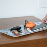 Birchwood tray on a wooden surface with keys, orange-strap watch, phone, pen, and black case; a hand reaches for items in Scandinavian Hygge style.
