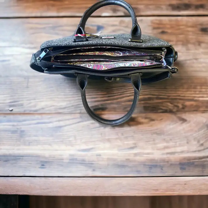 A Harris Tweed Double Handle Tote in grey rests on a wooden table, its open compartments displaying a vibrant patterned interior lining.