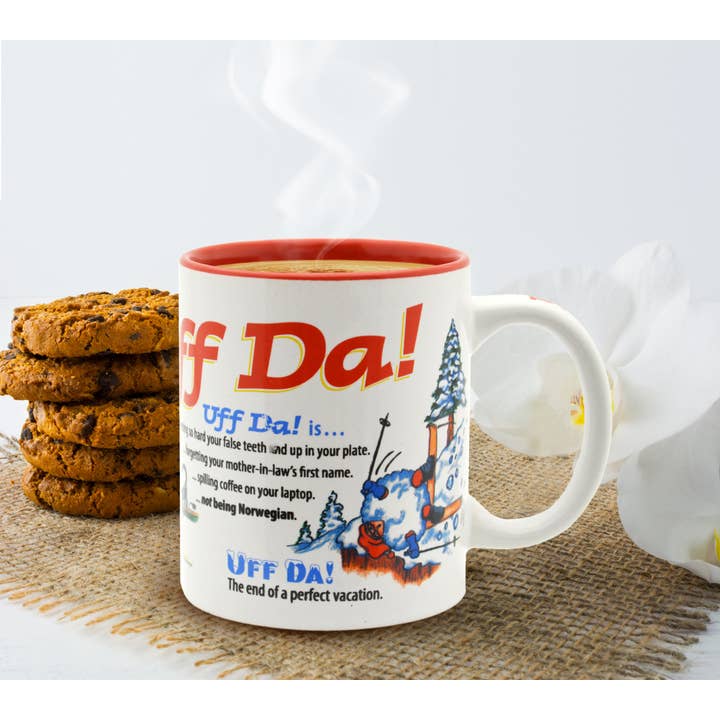 A steaming 12 oz mug with a red interior features Uff Da!—a nod to Minnesotas Scandinavian slang—and humorous sayings alongside a house illustration. Nearby, three cookies rest on a textured surface, surrounded by delicate white flowers in the background.