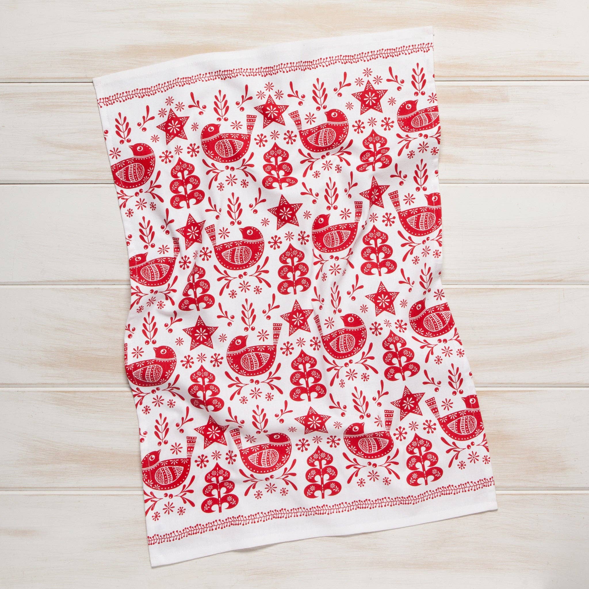 The Tea Towel: Snowbird Dishtowel, made from 100% cotton, features a charming pattern of birds, trees, and stars. With its red and white design, it adds a touch of charm to any kitchen against a light wooden background.