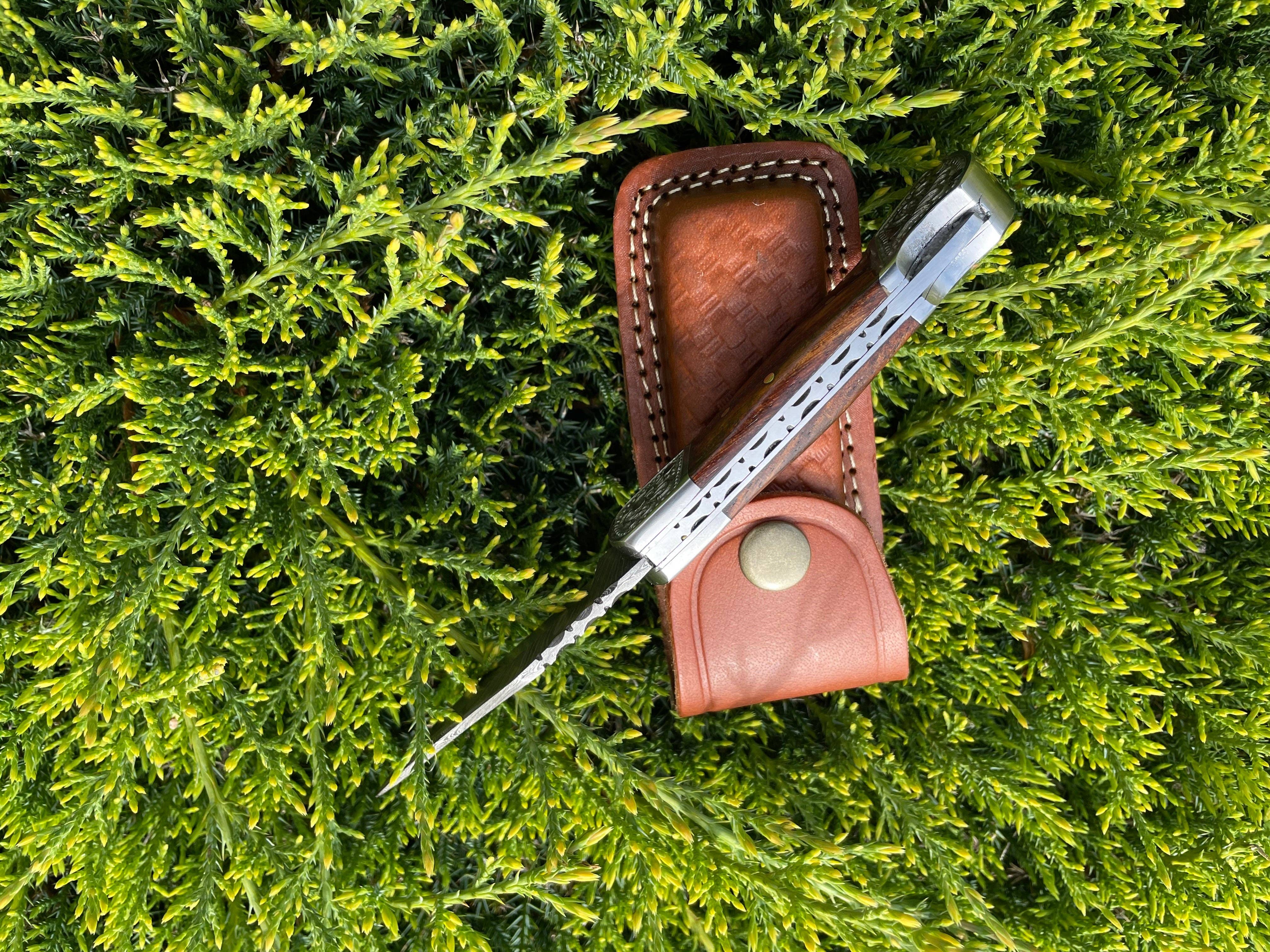 The Knife: Pocket Knife Walnut Wood 6.5'' Damascus Steel, partially open with its patterned blade and walnut wood handle, rests on a brown leather sheath atop dense green foliage.