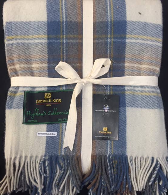 A Stewart Muted Blue Deluxe Highland Blanket, featuring blue, white, and beige plaid, is folded with a white ribbon and tags from Patrick King’s Highland Collection.