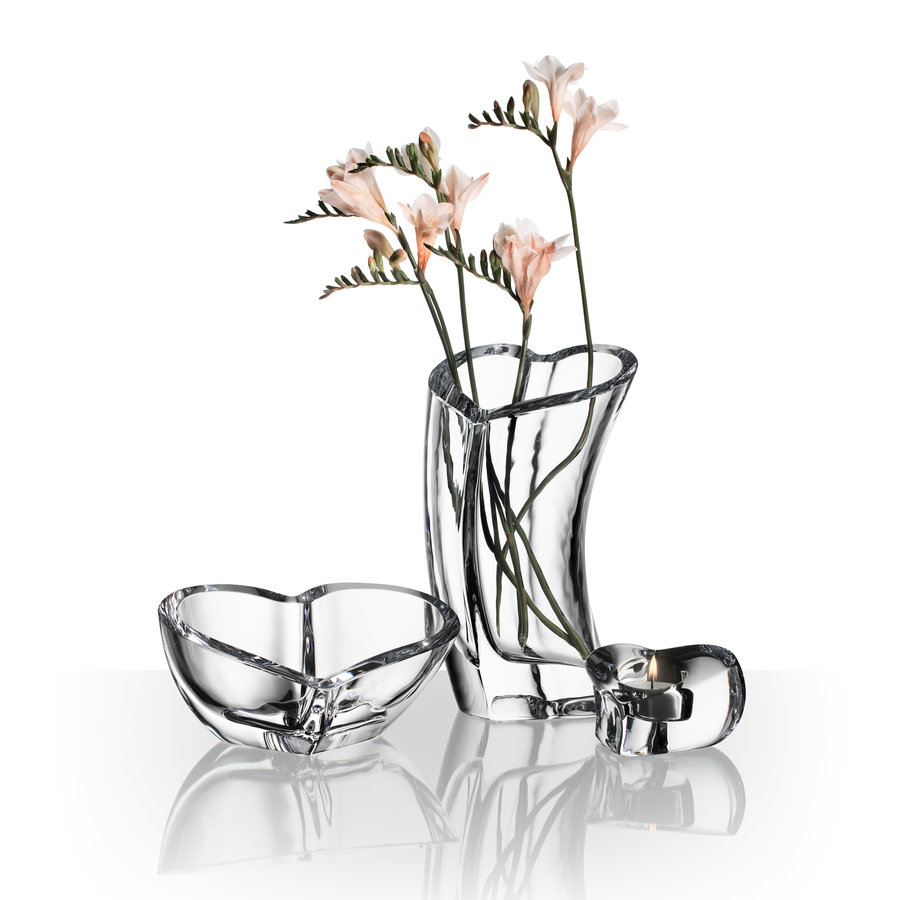Three clear glass vessels—a heart-shaped bowl, an Orrefors Valentino Votive Clear Candle Holder by Martti Rykonen, and a small round bowl—are arranged on a white reflective surface.