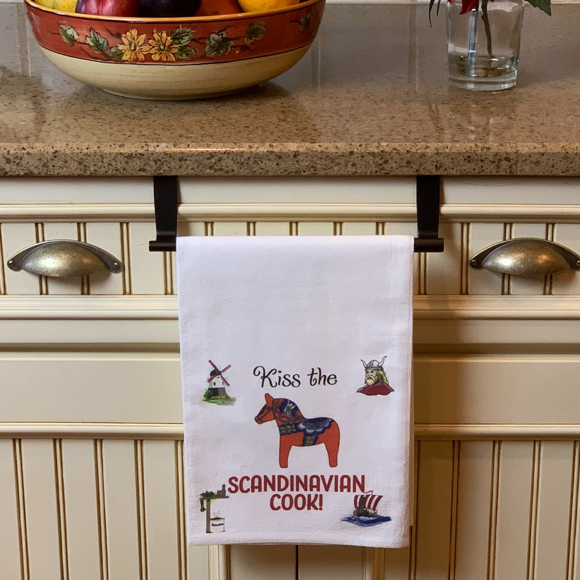 A Tea Towerl: Kiss the Scandinavian Cook Kitchen Gift Decorative Print hangs on a cabinet handle. This charming towel showcases illustrations of a windmill, Dala horse, Viking helmet, and ship, making it the ideal gift for any Scandinavian-themed kitchen.