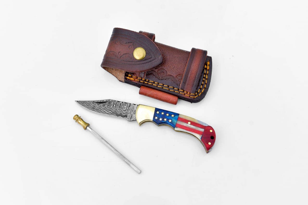 The Knife: Damascus Steel Pocket Knife Titan Patriot features an American flag handle, patterned blade, leather sheath, and sharpening rod—an eye-catching addition to your EDC, shown displayed on a white surface.