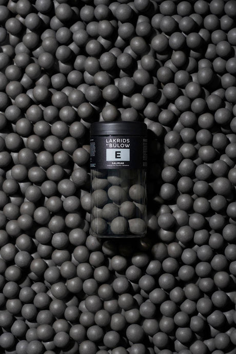 A jar labeled “E” is placed on a surface covered with round, matte black Nordic licorice balls arranged densely as a textured background.