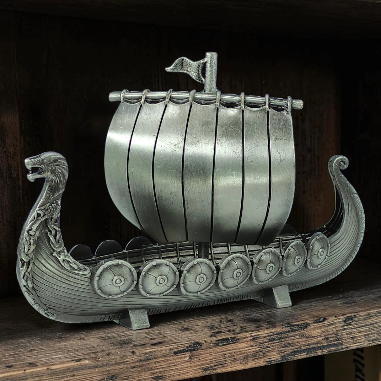 Display your Scandinavian heritage with the Faire - Scandinavian Specialties Figurine: Norwegian Viking Ship with Sail Pewter 5″x3″ Table Decor—a finely crafted pewter model featuring a dragon-shaped prow, striped sail, and side shields, beautifully presented on your shelf.