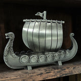 Display your Scandinavian heritage with the Faire - Scandinavian Specialties Figurine: Norwegian Viking Ship with Sail Pewter 5″x3″ Table Decor—a finely crafted pewter model featuring a dragon-shaped prow, striped sail, and side shields, beautifully presented on your shelf.