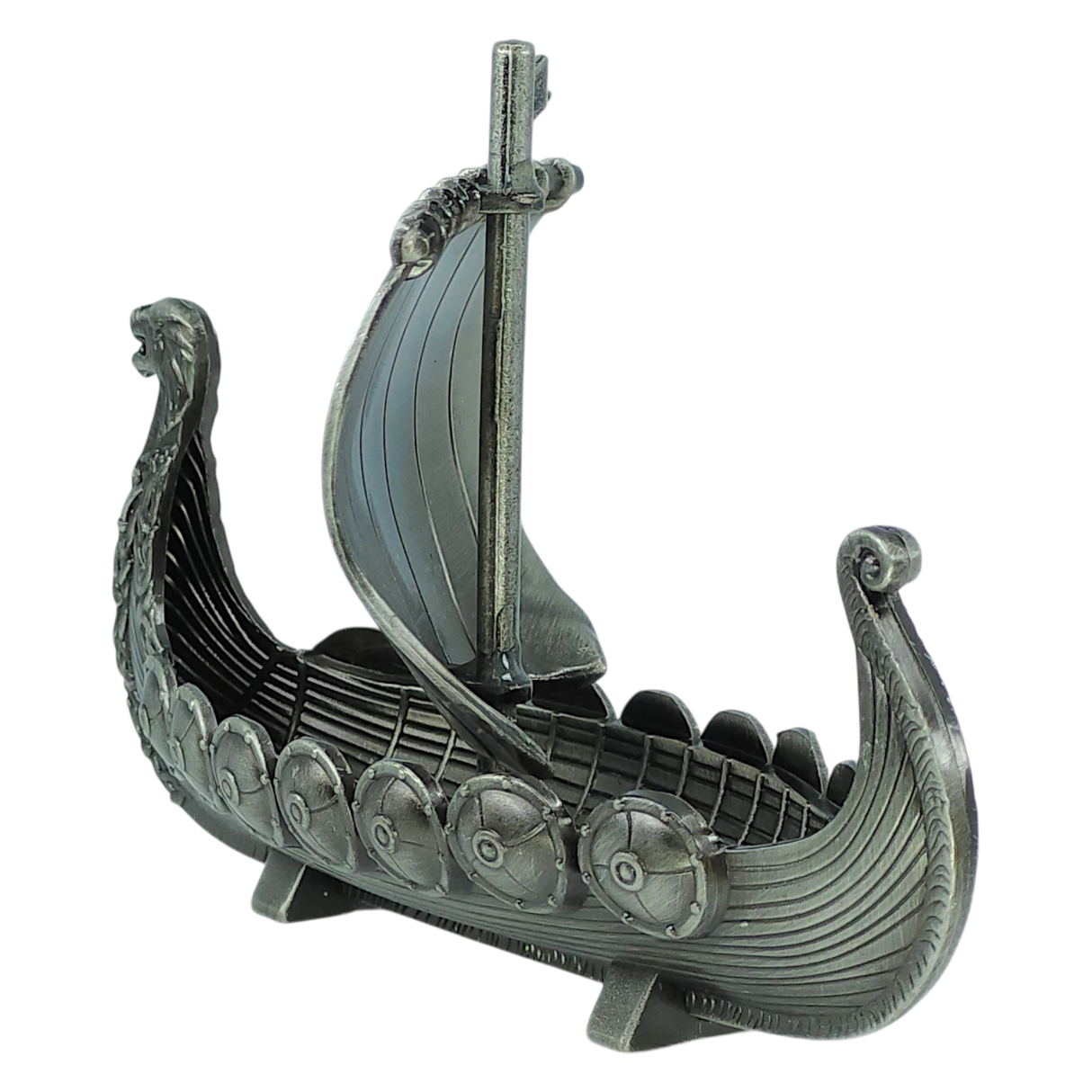The Faire - Scandinavian Specialties Figurine: Norwegian Viking Ship with Sail Pewter 5″x3″ Table Decor features a silver-toned model of a Viking longship, complete with an intricately detailed dragon head prow, shield decorations lining the sides, and a single mast topped with a sail. Celebrating Scandinavian heritage, this pewter decor piece is showcased on a plain white background.