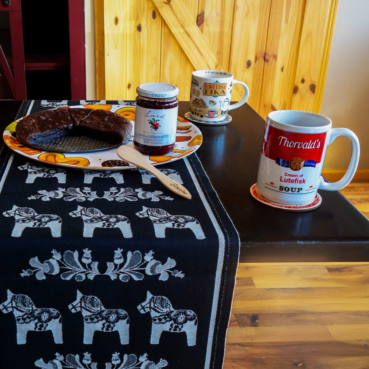 A wooden table holds a cake on a plate, two jars of jam, and Thorvalds Cream of Lutefisk Soup novelty mugs (15oz). A black table runner with white horse patterns enhances the rustic charm, alongside the wooden wall and door in the background.