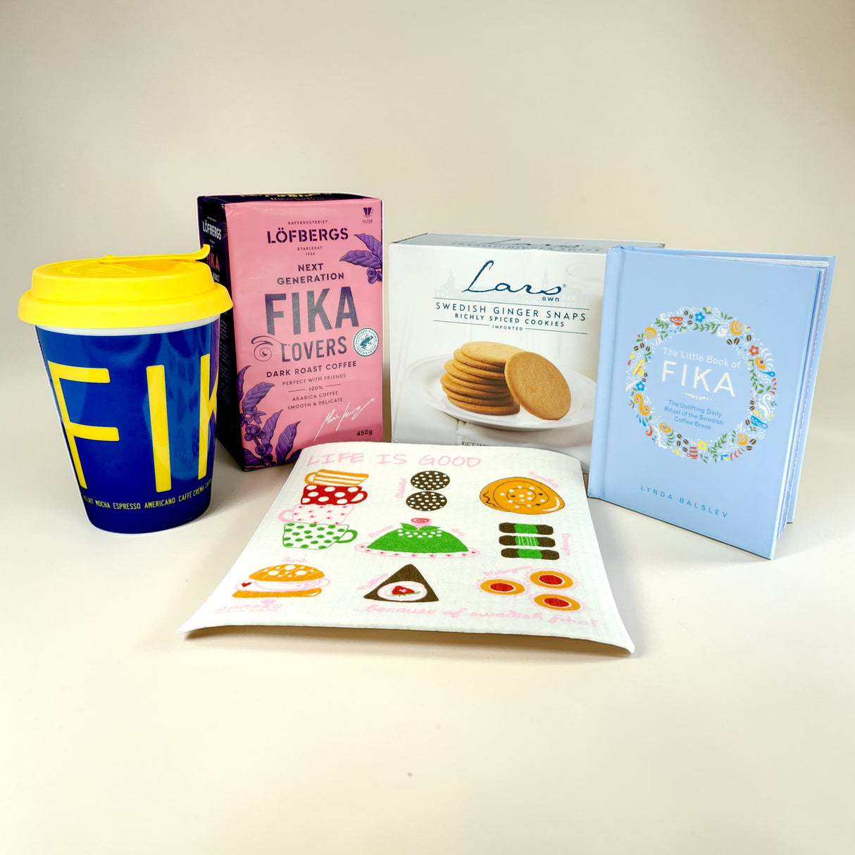 The Fika Starter Pack includes a yellow and blue travel mug, Löfbergs Dark Roast coffee, ginger snaps, a book, and a dessert-illustrated napkin—everything you need for an authentic Swedish coffee break.