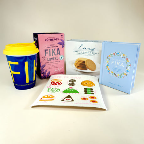 The Fika Starter Pack includes a yellow and blue travel mug, Löfbergs Dark Roast coffee, ginger snaps, a book, and a dessert-illustrated napkin—everything you need for an authentic Swedish coffee break.