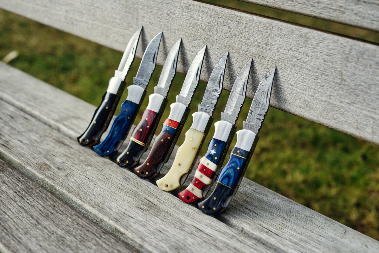 Seven pocket knives are lined up on a wooden bench outdoors, featuring unique decorative handles including a stunning Red & Black Wood Handle Genuine Damascus Steel Folding Knife.