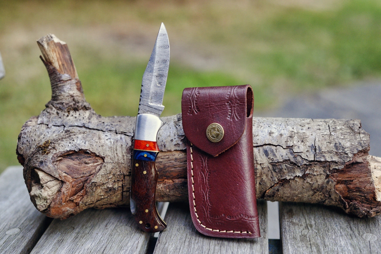 A Red & Blue Wood Handle Genuine Damascus Folding Knife is propped against a log on a wooden surface, next to its brown leather sheath with button closure, with grass visible in the background.