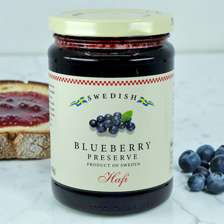 A jar of Swedish blueberry preserves on a marble surface, with wild blueberries and a slice of bread spread with the preserve in the background.