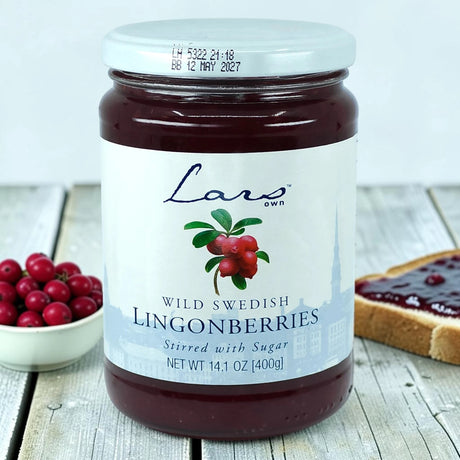 A jar of Swedish lingonberry jam on a white wooden table next to a bowl of berries and bread with Scandinavian preserves spread.