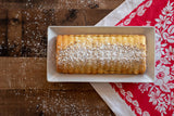 Rectangular pound cake with powdered sugar on a white plate, shown on a wooden table covered by a red floral cloth. Scandinavian almond cake pan design visible.