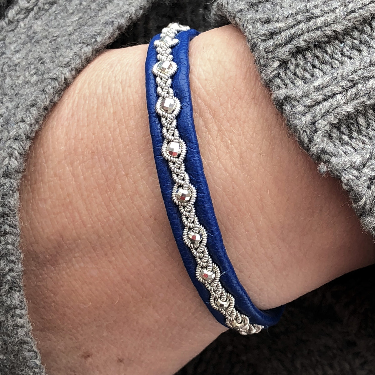 The Sami Frida Navy bracelet, hand-braided and adorned with silver embellishments, complements a cozy gray knitted sweater on a persons wrist.