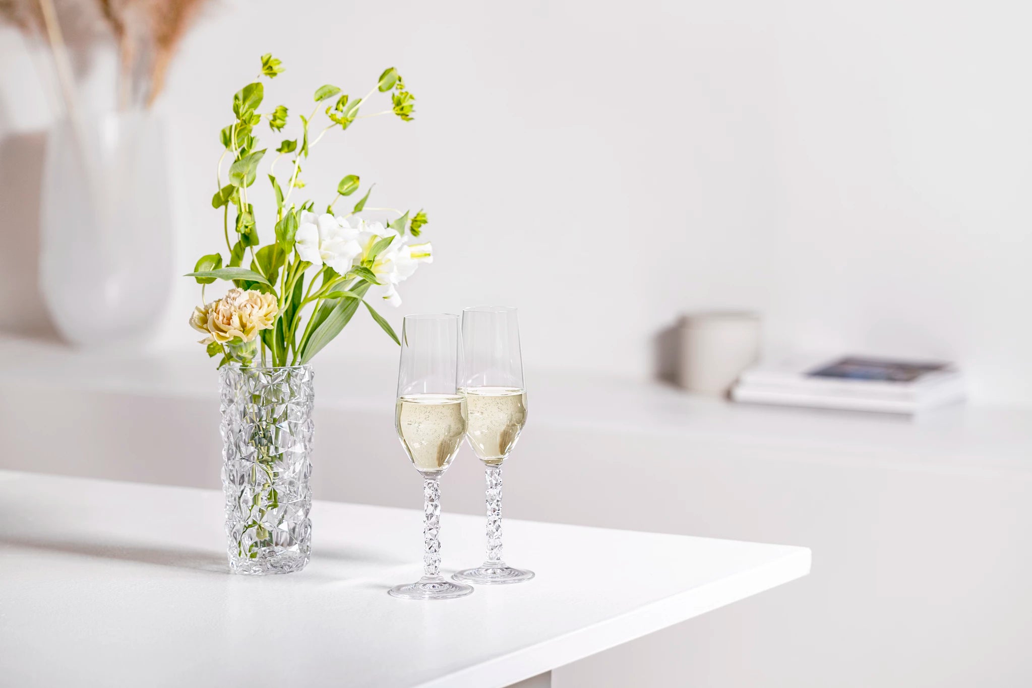 Crystal vase with green and white flowers is next to the Orrefors: Carat 3 Piece Gift Set champagne flutes on a white table.