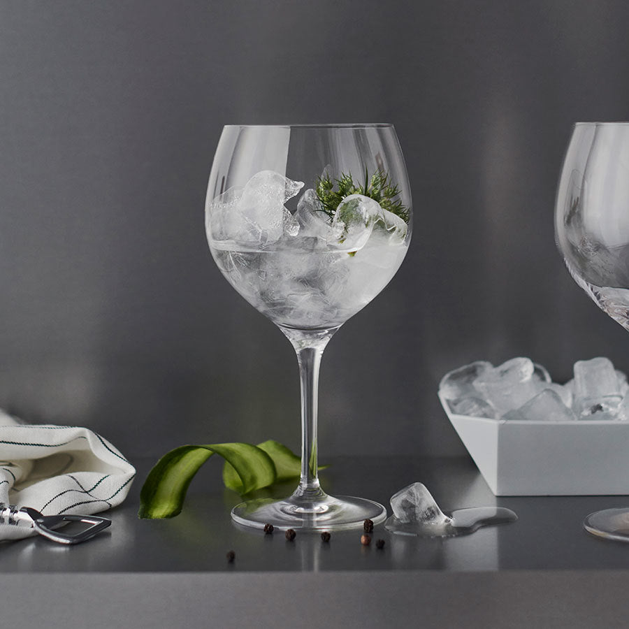 A gin and tonic in an Orrefors 22oz Gin and Tonic Glass with ice, lime, and herbs captures Barcelonas drinking vibe. A towel, peeler, tray, and scattered ice enhance the scene on a reflective surface.