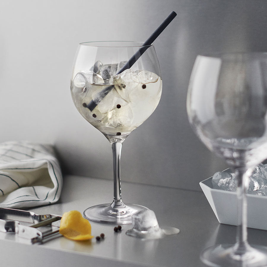 On a table sits an Orrefors Gin and Tonic Glass filled with a refreshing drink, ice cubes clinking against a black straw. Nearby, metal tongs rest beside a lemon peel, capturing the essence of Barcelona drinking culture.